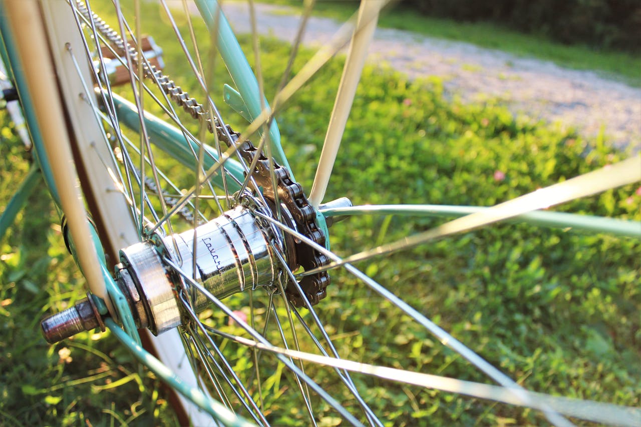 Close up photo of the wheel of a light blue and white bicycle showing the psokes coming off the hub and the bicycle chain. The bicycle sits in the grass next to a blurred gravel trail.