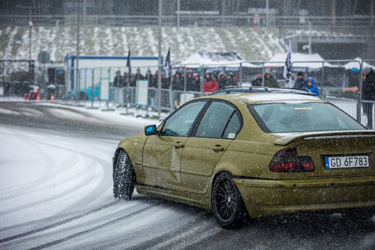 A mustard yellow car drifts across the snow along a track with spectators watching from beyond a chainlink fence.