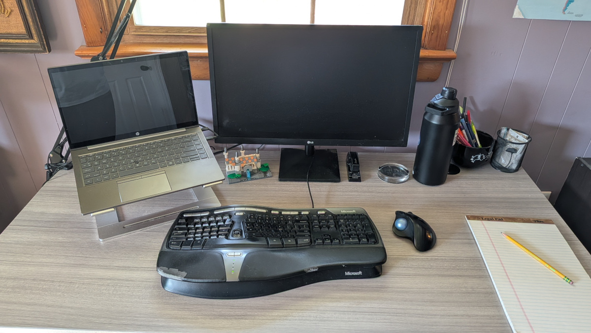 A gray office desk, freshly cleaned, sporting a keyboard and trackball, laptop on a stand, monitor, water ball, desk lamp, cup of pens, new legal pad with a pencil, and a LEGO Rivendell.