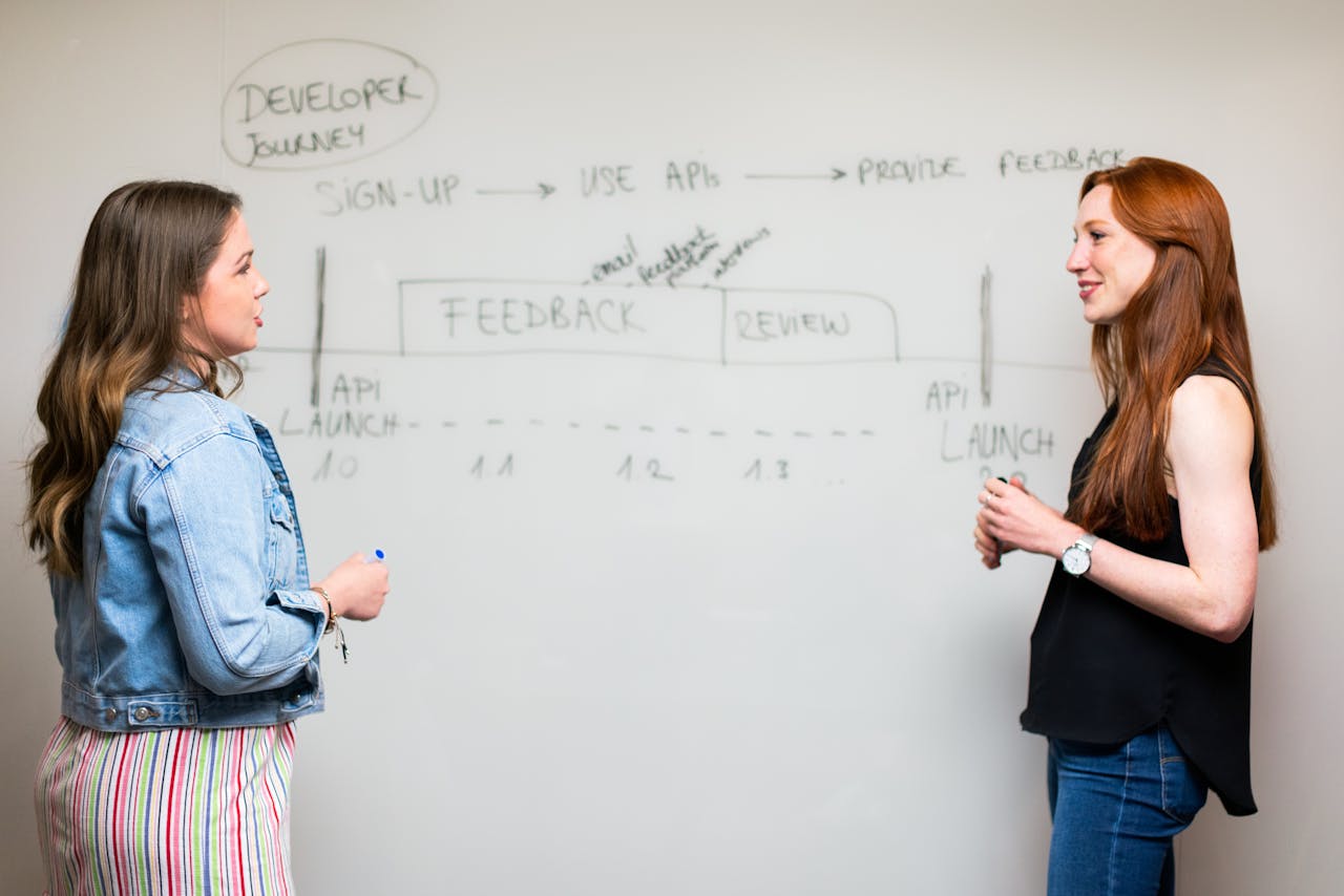 Two women stand on opposite ends of a white board. One holds a marker while explaining a diagram to the other.