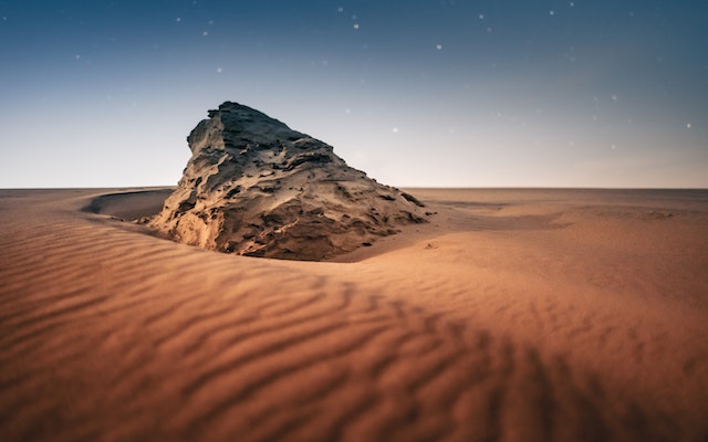 A craggy rock juts out of windswept desert sands under a star studded twilight sky with the last remnants of sunlight peeking over the horizon.