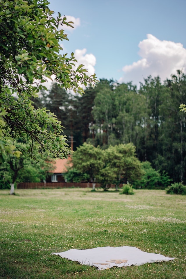 A white blanket sits on a well tended green lawn. Trees surround the park-like atmosphere. A few puffy white clouds float in an otherwise blue sky.