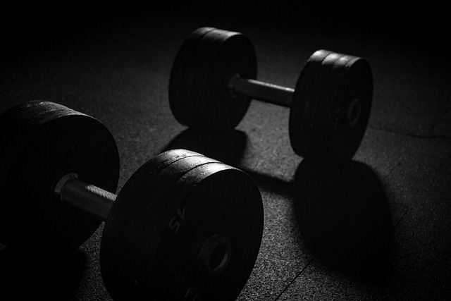 A pair of black dumbbells rest on a dark floor. A beam of light comes from somewhere up above.