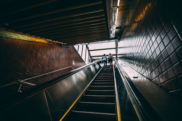 Looking up from the bottom of a subway's escalator ascending to street level. Two people ride at the top.