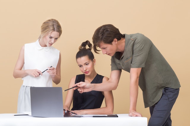 A woman sits at a desk with a laptop computer. Another woman stands to the person's right while a man to the person's left leans over the desk and uses a pen to point at something on the laptop screen.