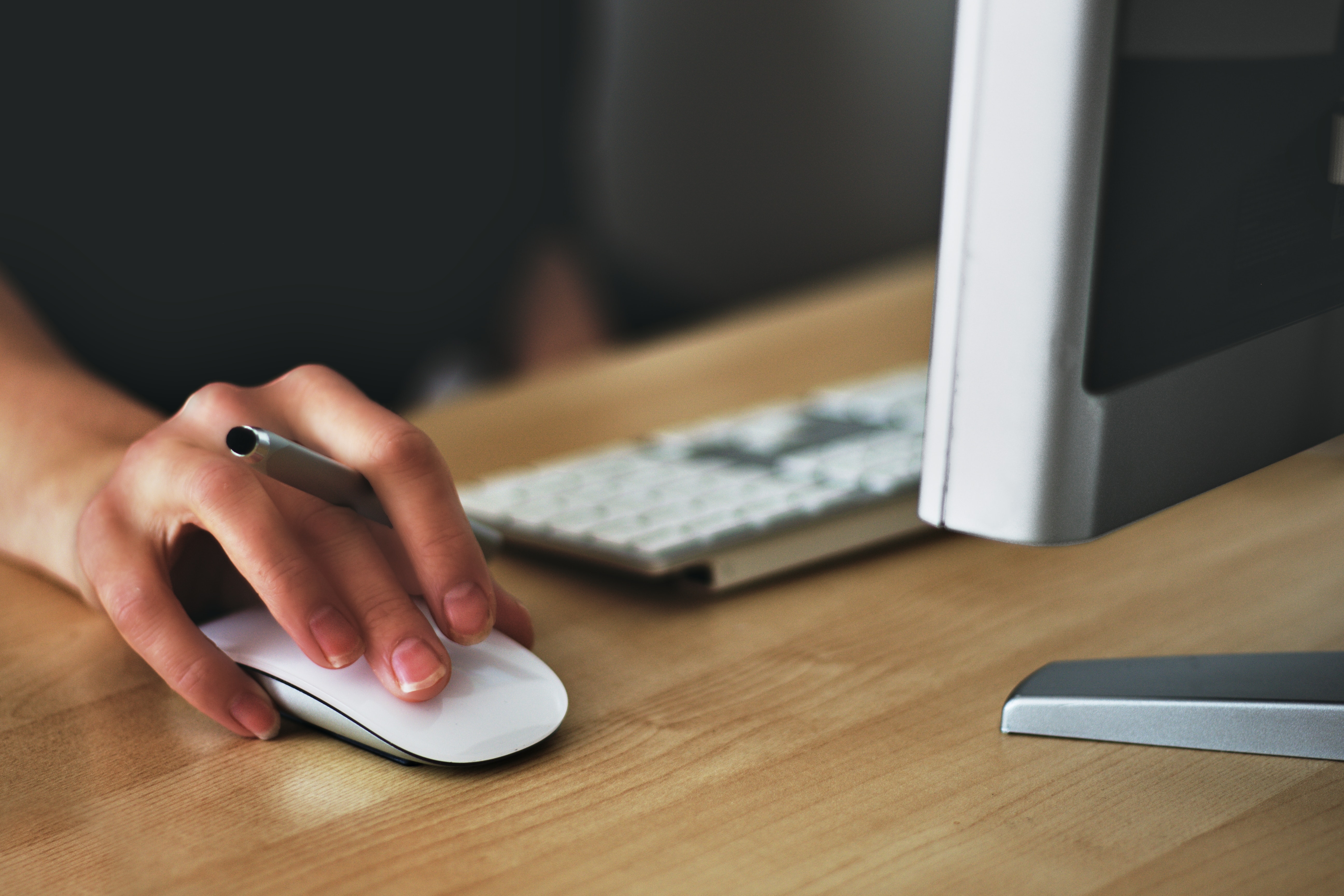 A person sitting at a desk with a monitor, keyboard, and mouse. The angle focuses on the hand holding a pen on top of the computer mouse.