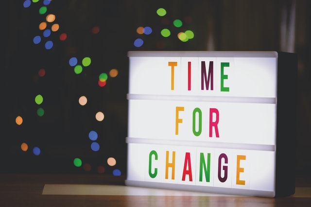 A white, lit up letter board uses multi colored letters to say Time for change. In the background is what looks like a very blurred lit up Christmas tree.