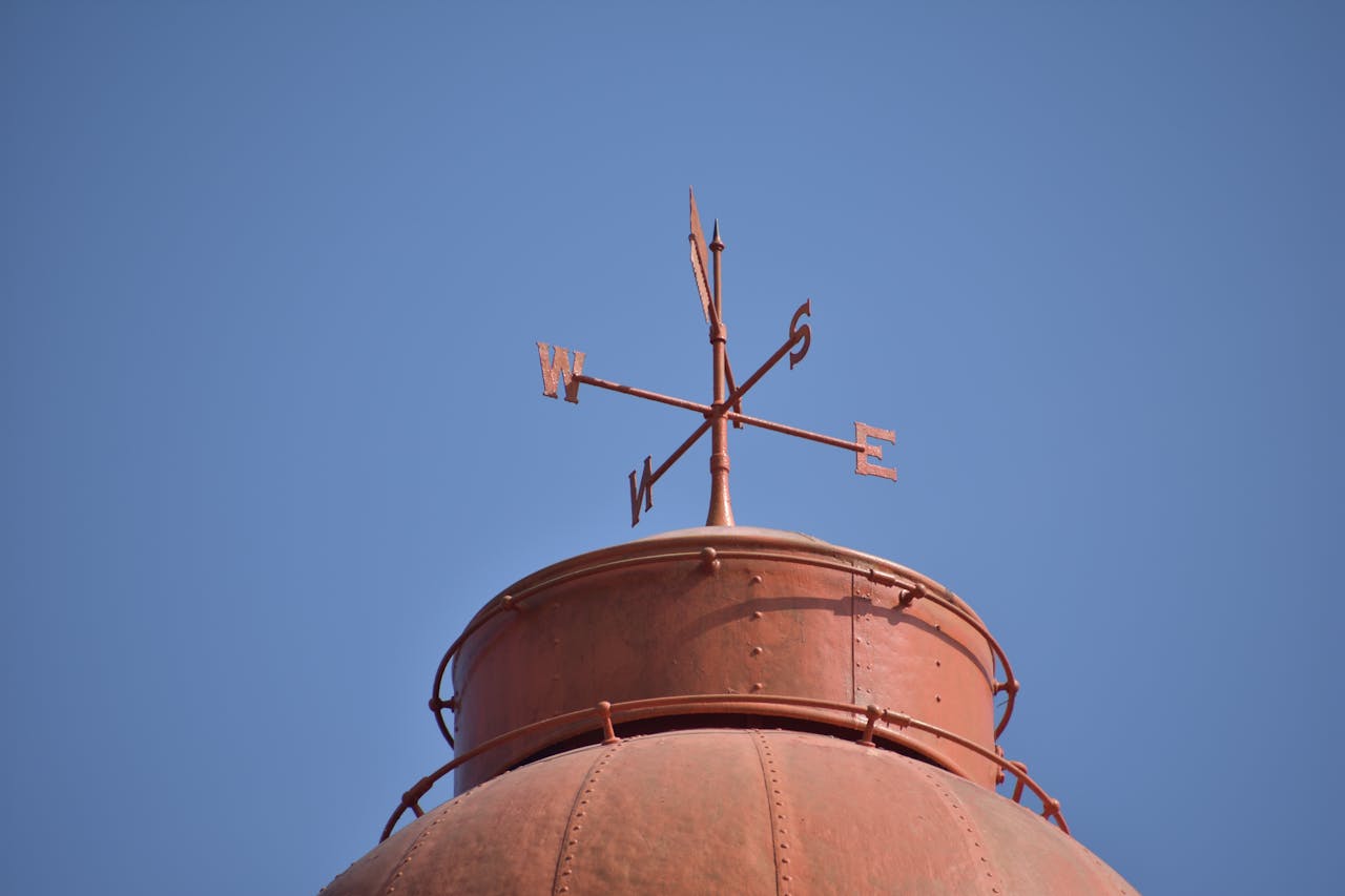 Looking up at a wweather vane atop a bronze rusted metal structure against a clear blue sky.